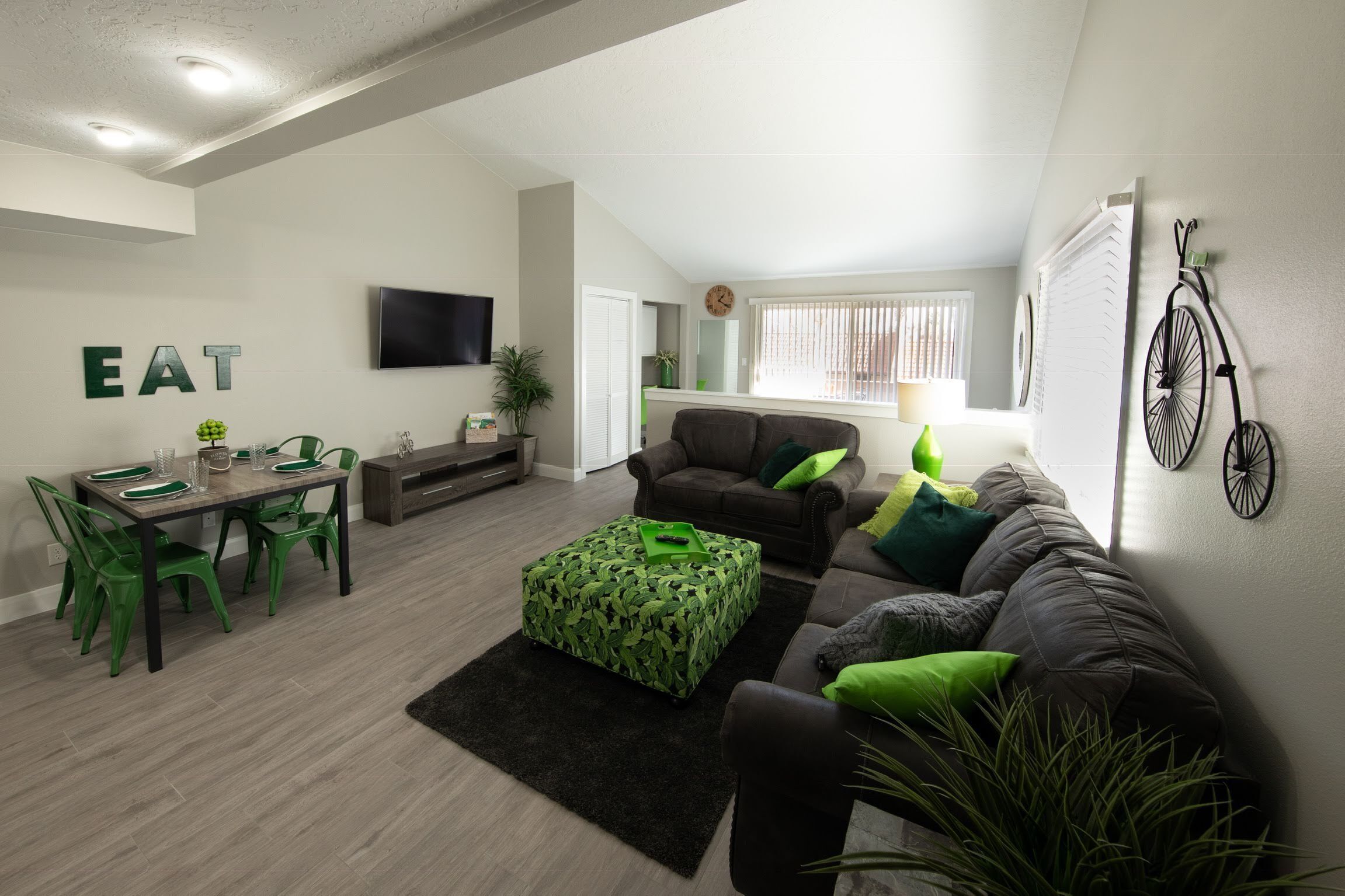 A modern living room with a dining area, a grey sofa, green cushions, and a flat-screen TV mounted on the wall.