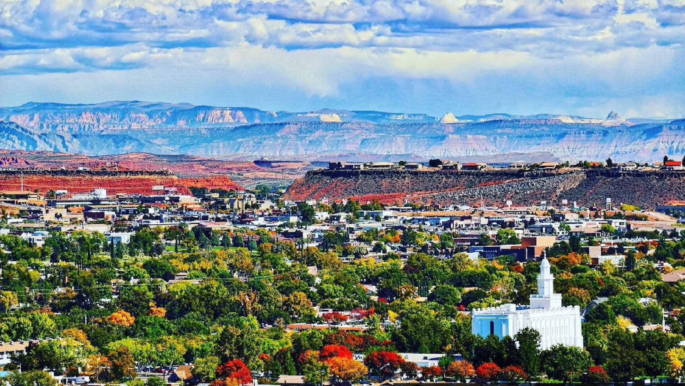A cityscape with a prominent white building, likely a government or historical structure, surrounded by dense greenery and a backdrop of red rock formations.