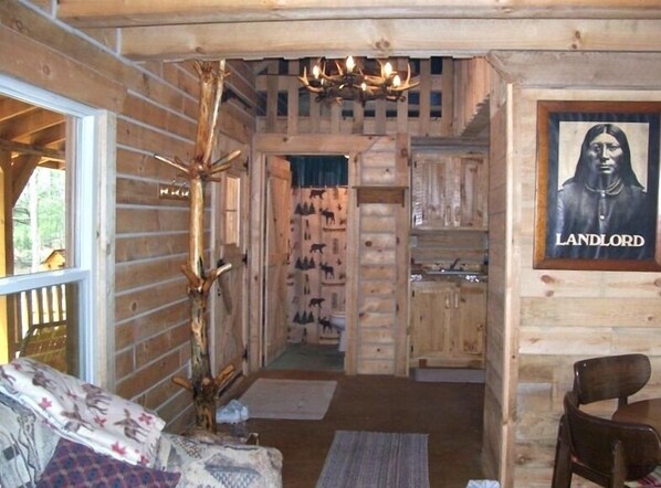 A log cabin interior with a wooden staircase, a chandelier, a framed picture labeled "LANDLORD," and a dining area with a table and chairs.