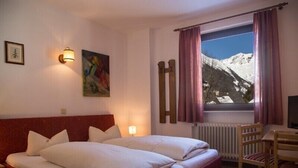 A hotel room with a bed, a desk, a chair, a window with a mountain view, and wall-mounted artwork. - Hotel Kärntnerhof Mallnitz (Mallnitz)