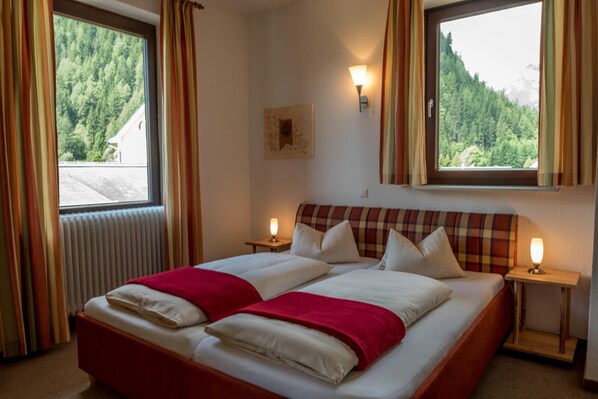 Uma cama de casal com um cobertor vermelho, duas mesas de cabeceira com luminárias, uma janela com cortinas e um quadro na parede.