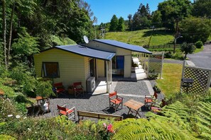 Un petit bâtiment d’un seul étage, doté d’un toit en pente, d’un patio avec des sièges extérieurs et d’un jardin planté de fougères.