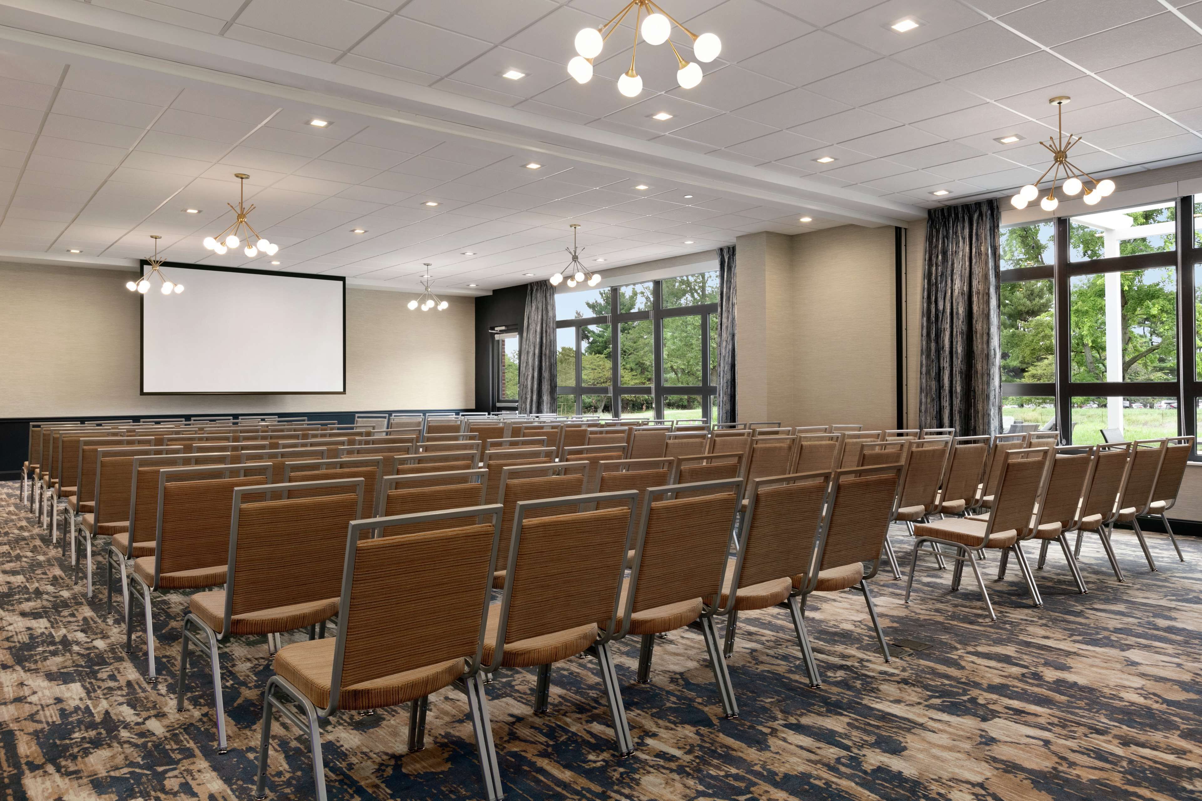 A conference room with rows of chairs, a large screen, and windows with a view of greenery.