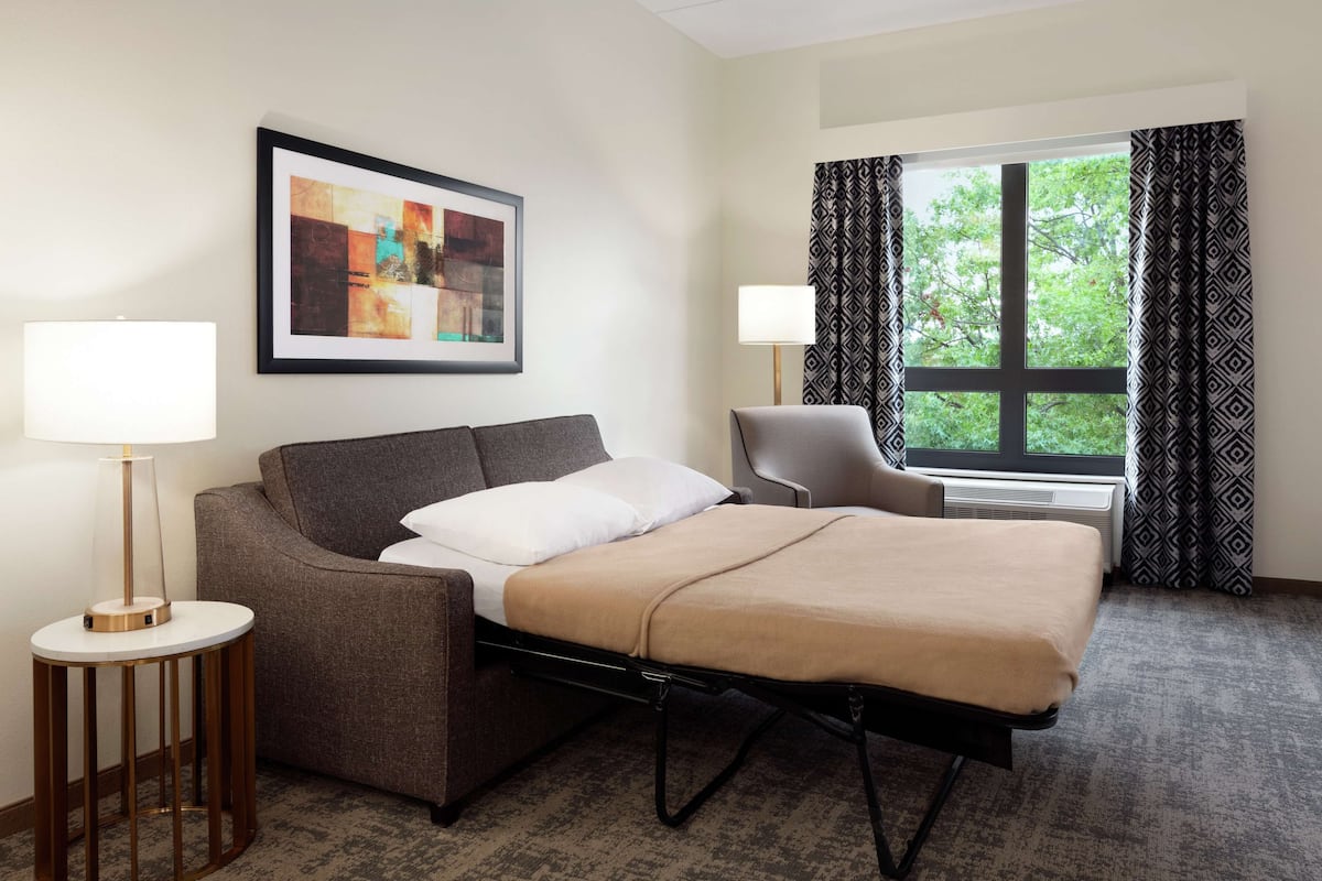 Ein Hotelzimmer mit Bett, Sessel, Lampe, einem gerahmten Kunstwerk und einem Fenster mit Vorhängen.