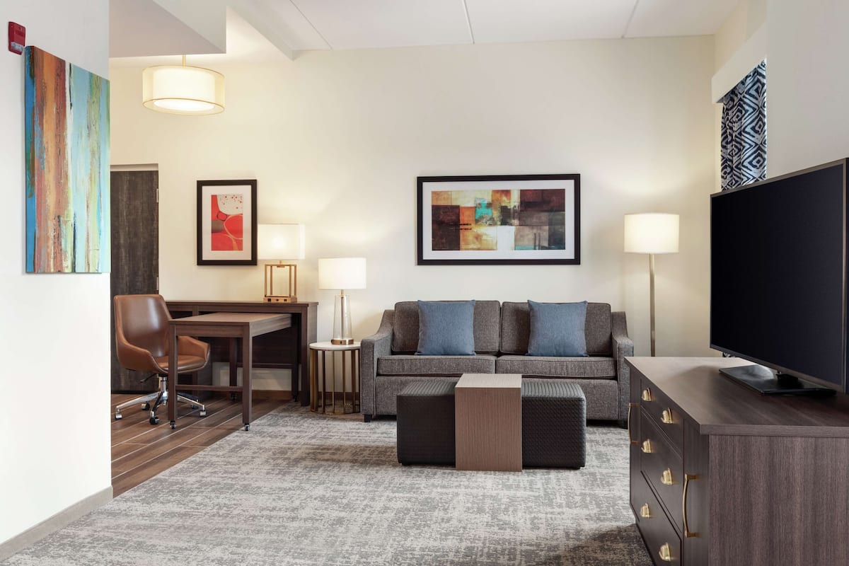 Ein modernes Hotelzimmer mit grauem Sofa, einem Flachbildfernseher, einem Holztisch und einem braunen Ledersessel.
