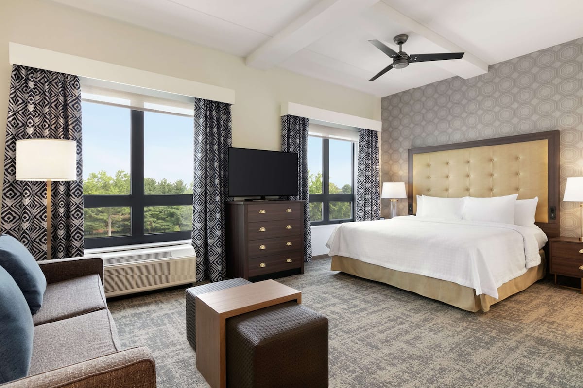 Ein Hotelzimmer mit einem großen Bett, einem Fernseher, einem Schreibtisch und einem Sessel. Vorhänge und ein Fenster mit Blick sind vorhanden.