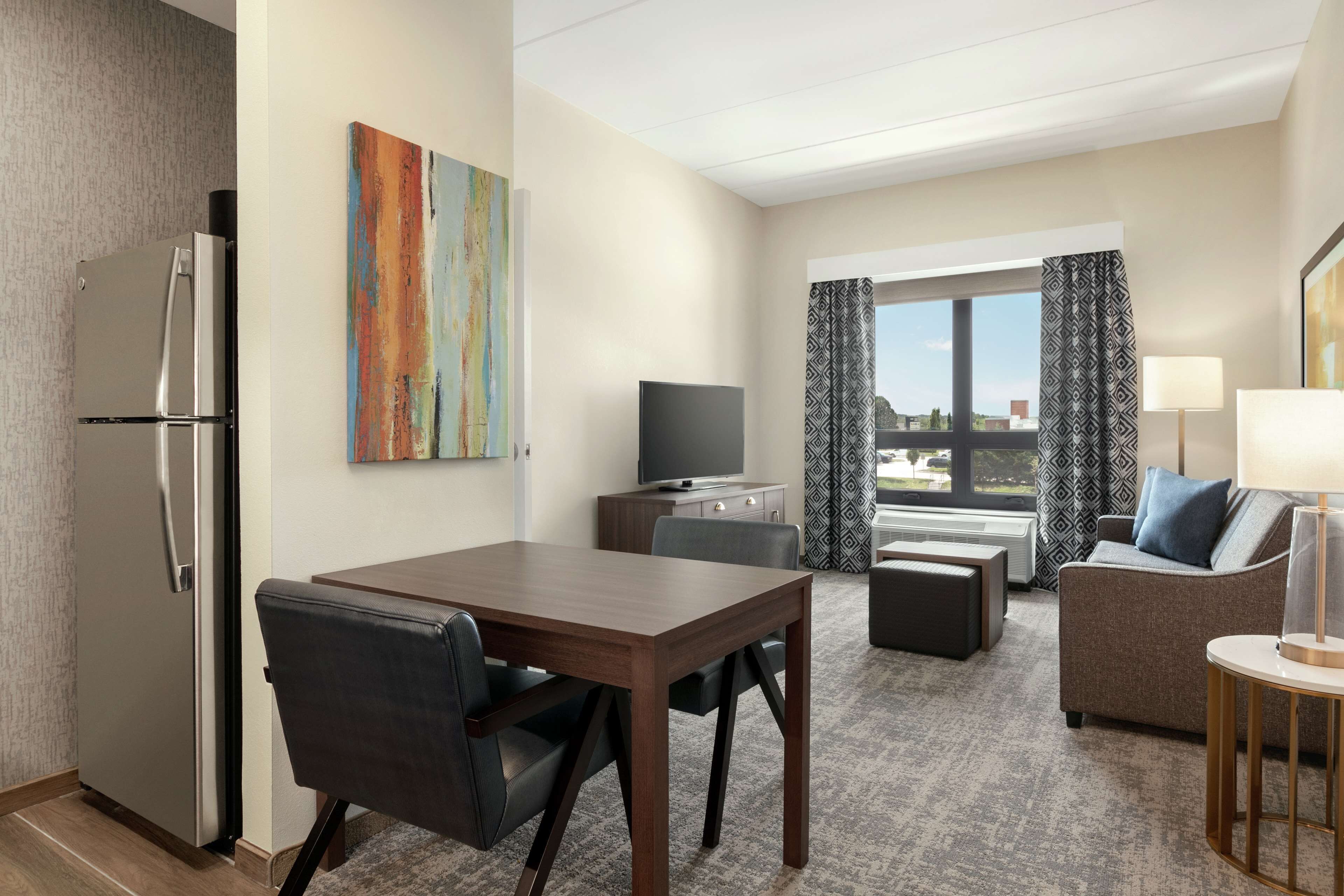 Habitación de hotel con una mesa de comedor, un sofá, un televisor, un refrigerador y una ventana con cortinas.