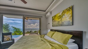 A bedroom with a large bed, a view of the outdoors, and a ceiling fan. - Exclusive Pool Oasis: Stunning Views! Sleeps 8, Nature's Seclusion (Puntarenas)
