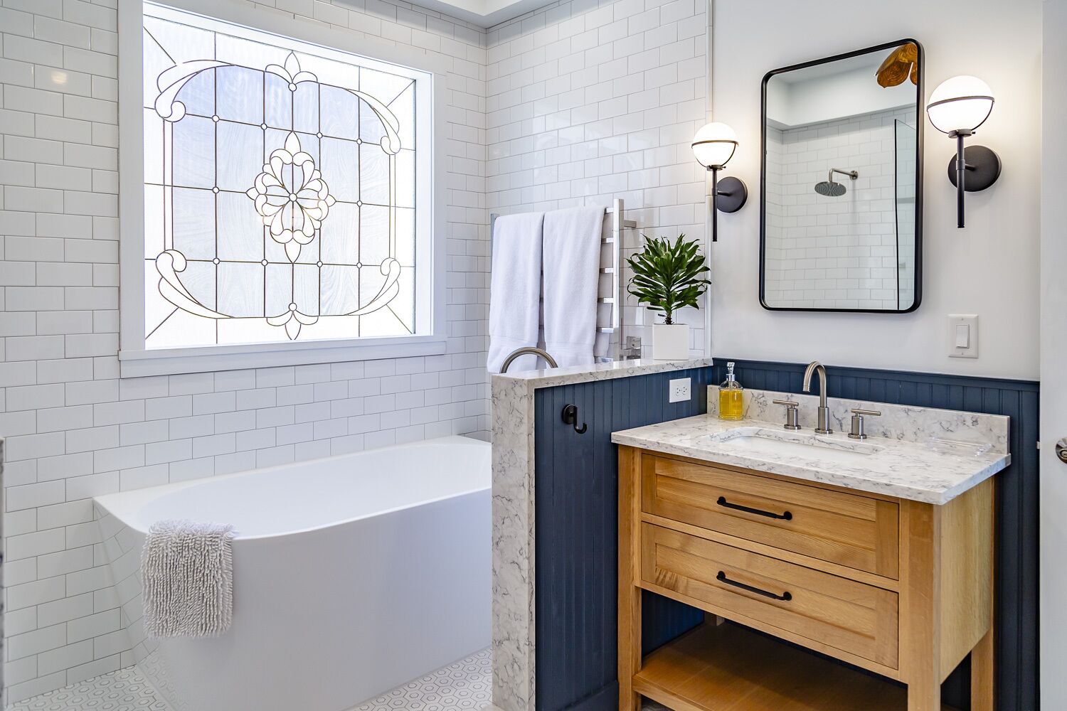 A modern bathroom with a white bathtub, a wooden vanity with a marble countertop, a large mirror, and a decorative window with a floral pattern.