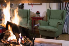 A living room with two green armchairs, a small wooden table with a vase of flowers, and a fireplace with visible flames. - Alpine Aria (Kalavrita)