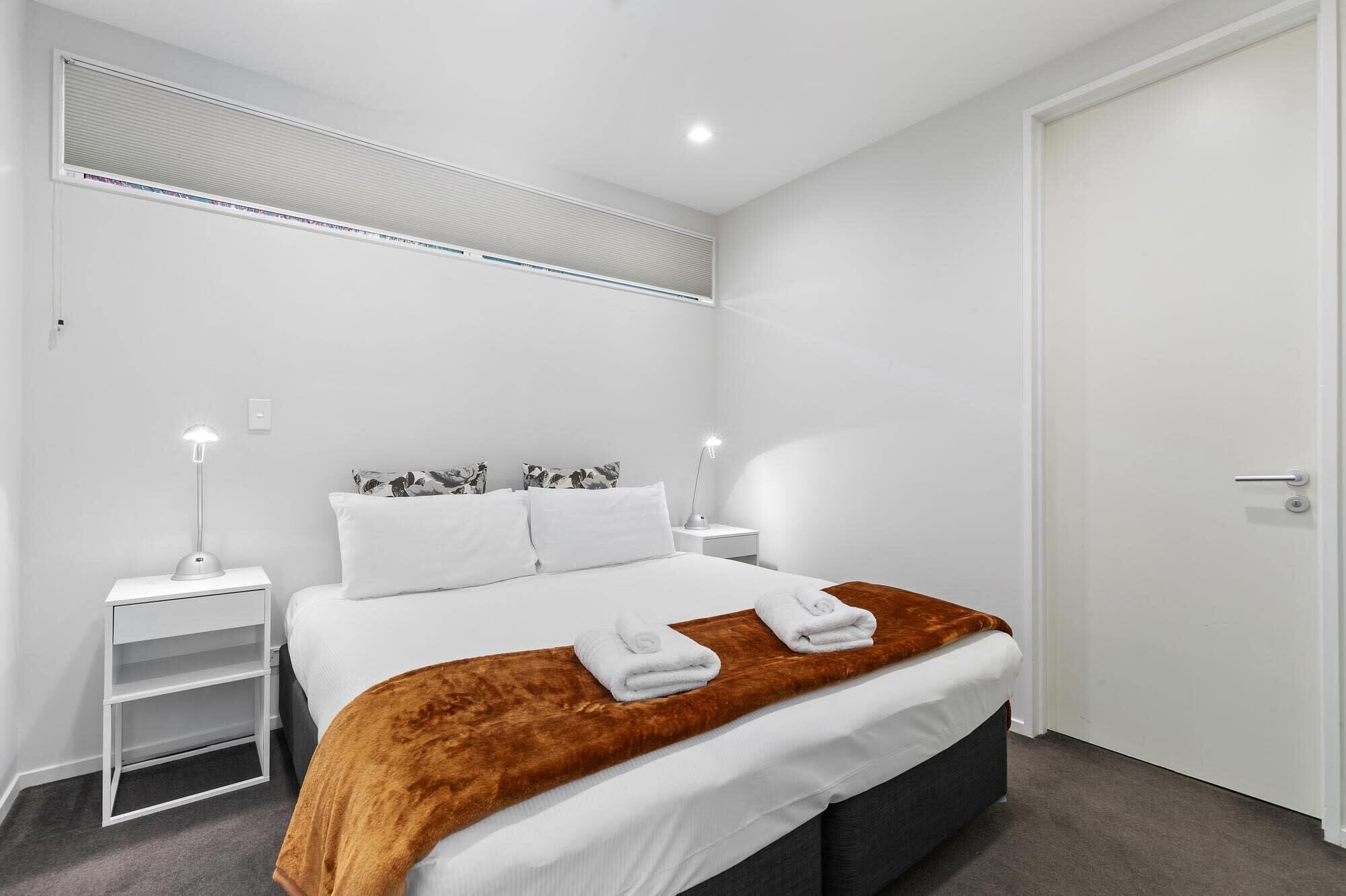 A modern bedroom with a bed, two bedside tables, a nightstand, a door, and a window with blinds.