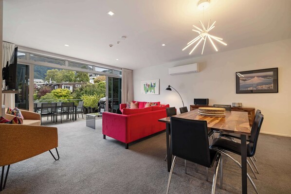 A modern living room with a red sofa, a dining table with chairs, and a large window overlooking a balcony with outdoor furniture. - Edgar Rise, 500m from Queenstown CBD (Queenstown Hill)