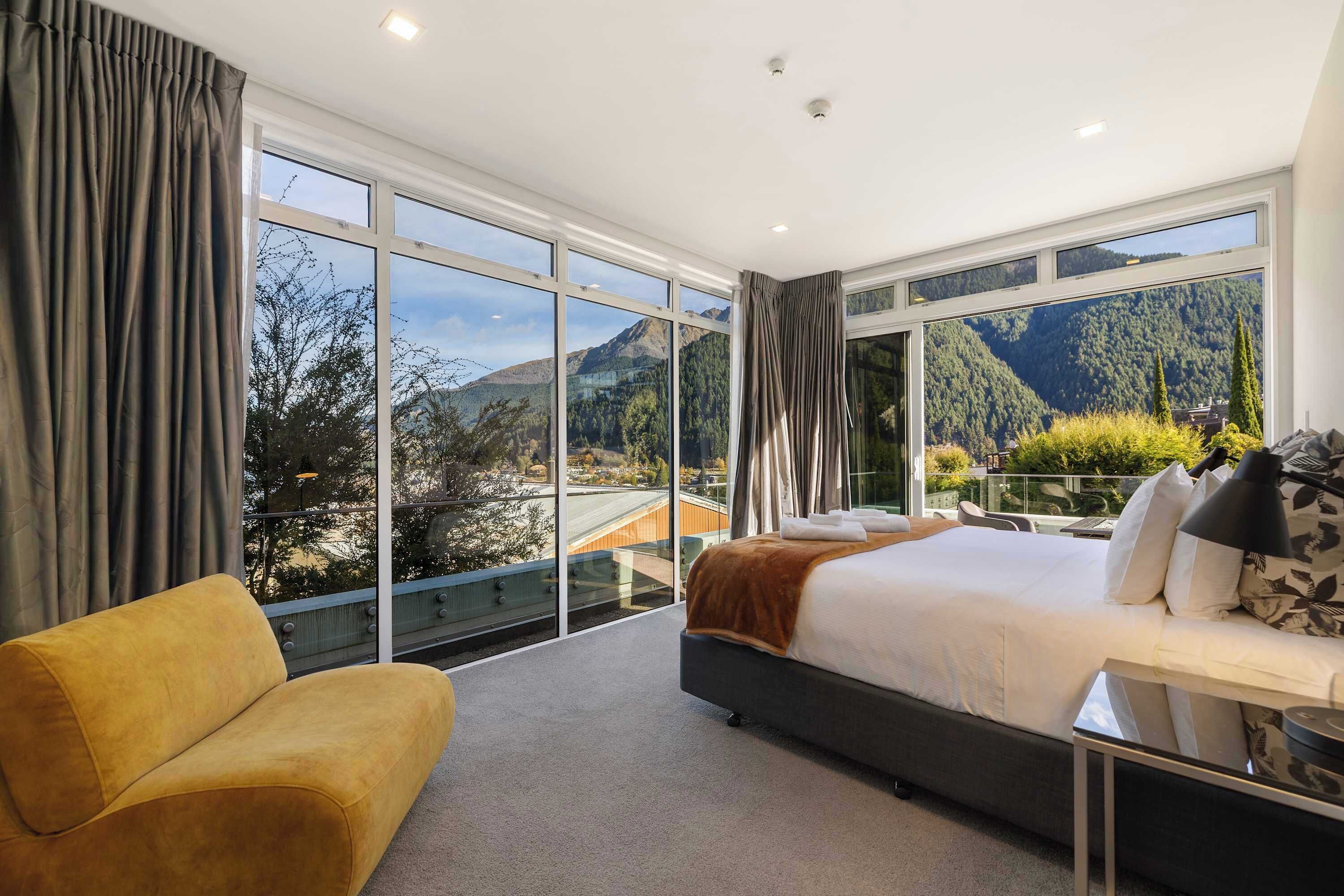 A modern bedroom with a large bed, a sofa, and a view of mountains through floor-to-ceiling windows.