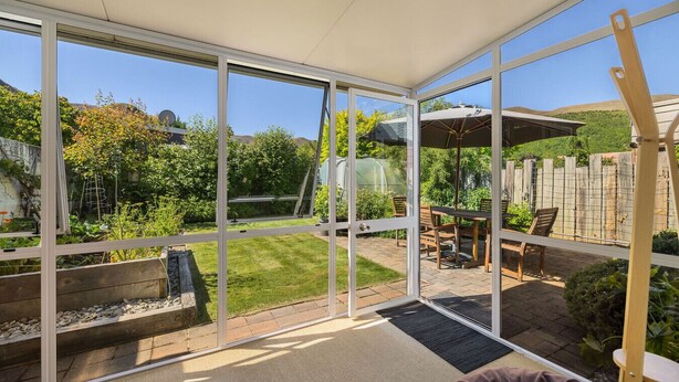 Une véranda dotée d’une porte vitrée, un espace patio avec une table et des chaises, et une vue sur un jardin et des collines.