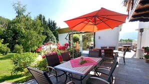 Een terras buiten met een tafel en stoelen, een rode parasol en uitzicht op de tuin.