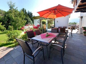 An outdoor patio with a table and chairs, a red umbrella, and a garden view. - Double room with extra bed - Pension Irlingerhof *** (Guggenberg)