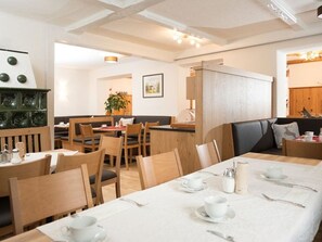 Een eetgedeelte met houten tafels en stoelen, een witte tafelkleed en uitzicht op een keuken.