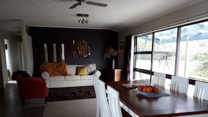 Un coin repas avec une table en bois et des chaises blanches, un canapé avec des coussins et une grande fenêtre donnant sur l’extérieur.