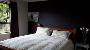 Une chambre avec un lit en bois, une fenêtre avec des rideaux, une table de chevet ornée d’un vase de fleurs et un tableau accroché au mur.
