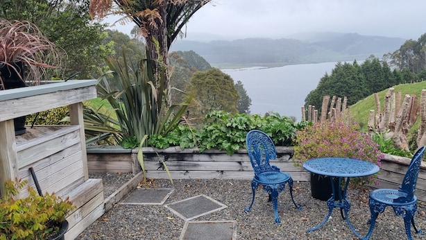 Une terrasse avec une table et des chaises, entourée de plantes et offrant une vue sur un lac et des collines.