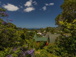 A house with a green roof and a small tower-like structure, surrounded by lush greenery and purple flowers. - Treetop Bach - Hahei Bach (Hahei)