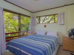 A bedroom with a bed, a nightstand, a dresser, a fan, and a window with curtains. - Treetop Bach - Hahei Bach (Hahei)