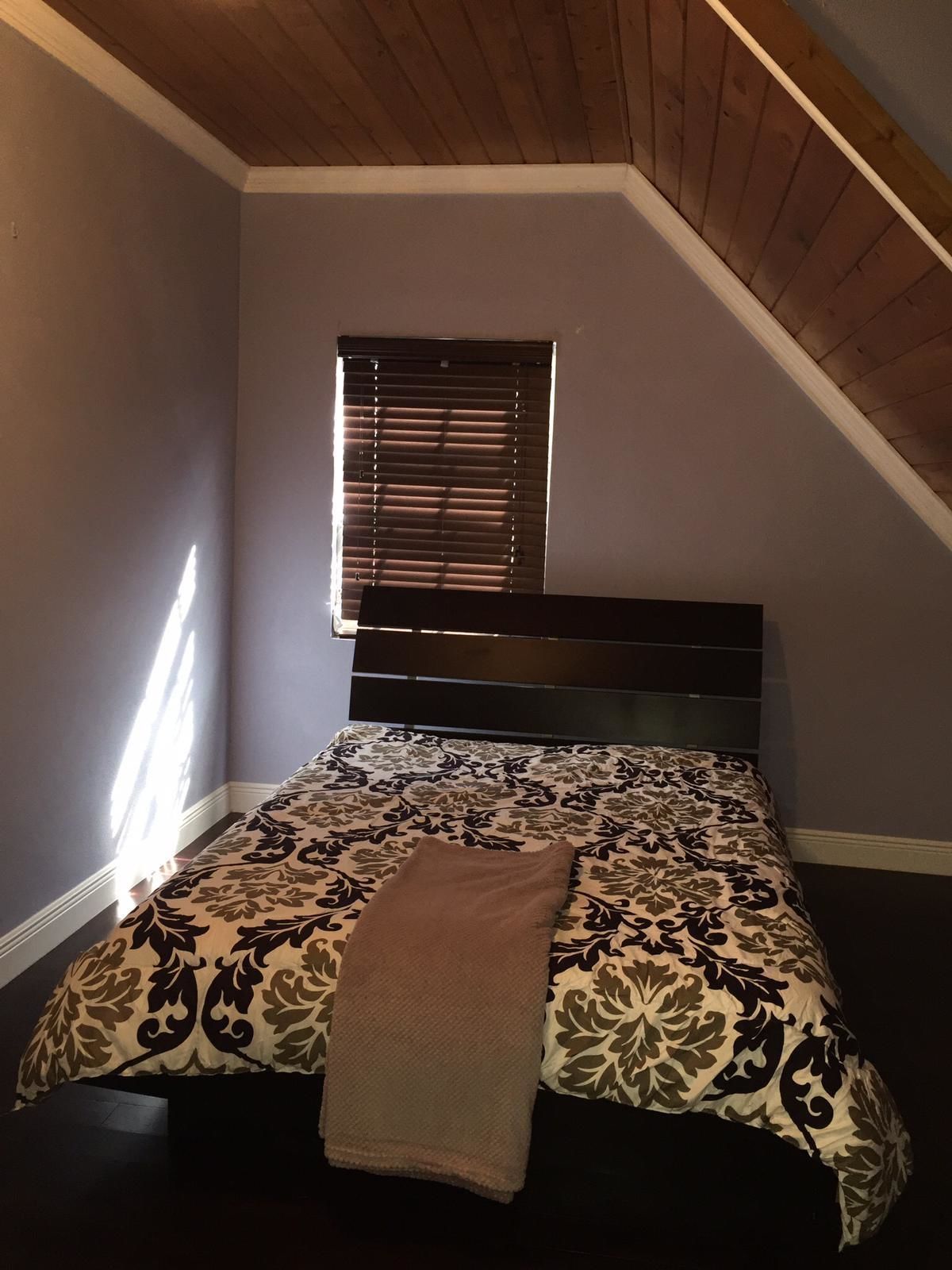 A bedroom with a sloped ceiling, a window with blinds, a bed with a patterned bedspread, and a brown throw blanket.