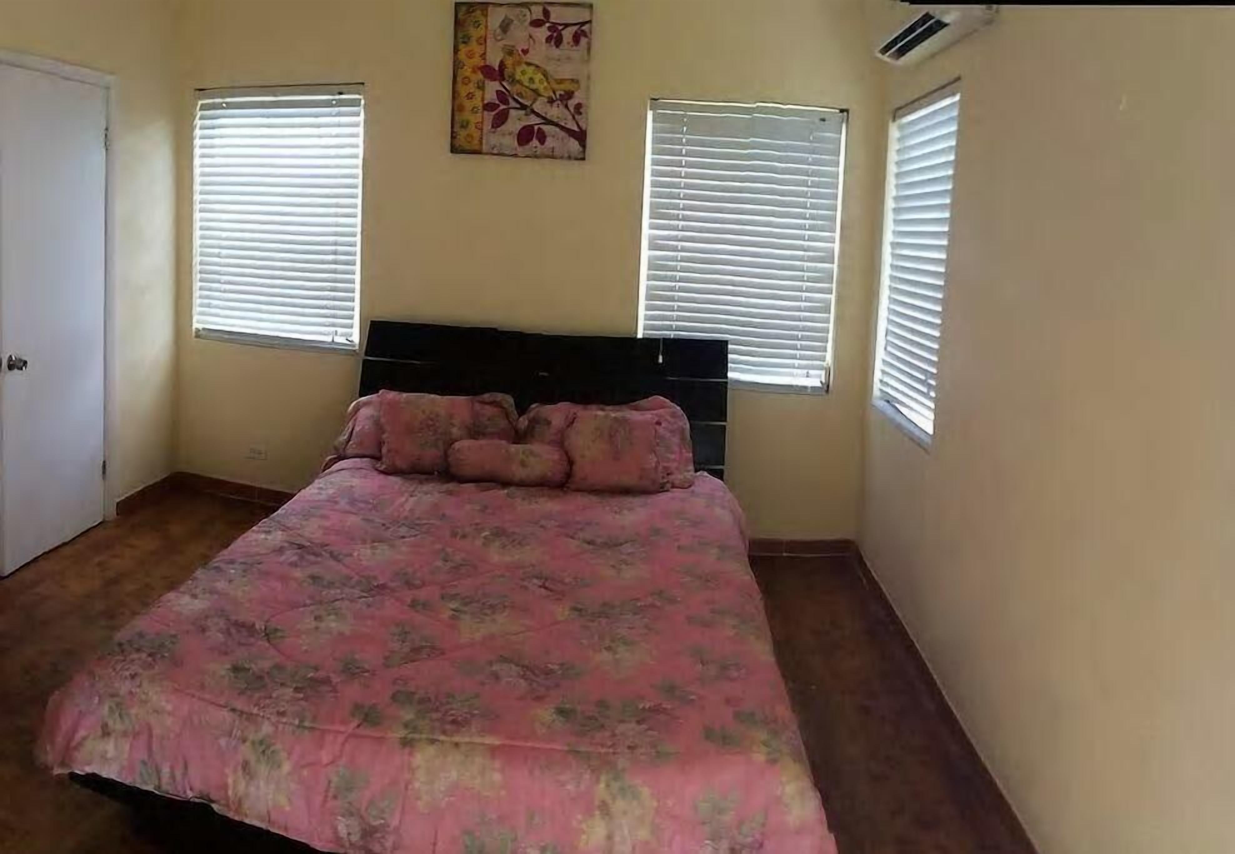 A bedroom with a bed, two windows with blinds, a door, and a wall-mounted air conditioner.