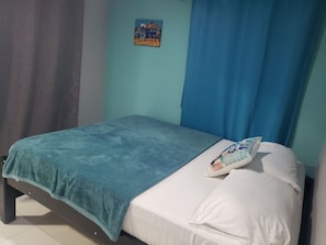A single bed with a teal blanket and white pillows, a small poster on the wall, and blue curtains. - Plaza del Mar (San Andrés)