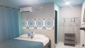 A bedroom with a bed, a wall-mounted air conditioner, a patterned wall decoration, a bedside table, and a hanging clothes rack. - Plaza del Mar (San Andrés)