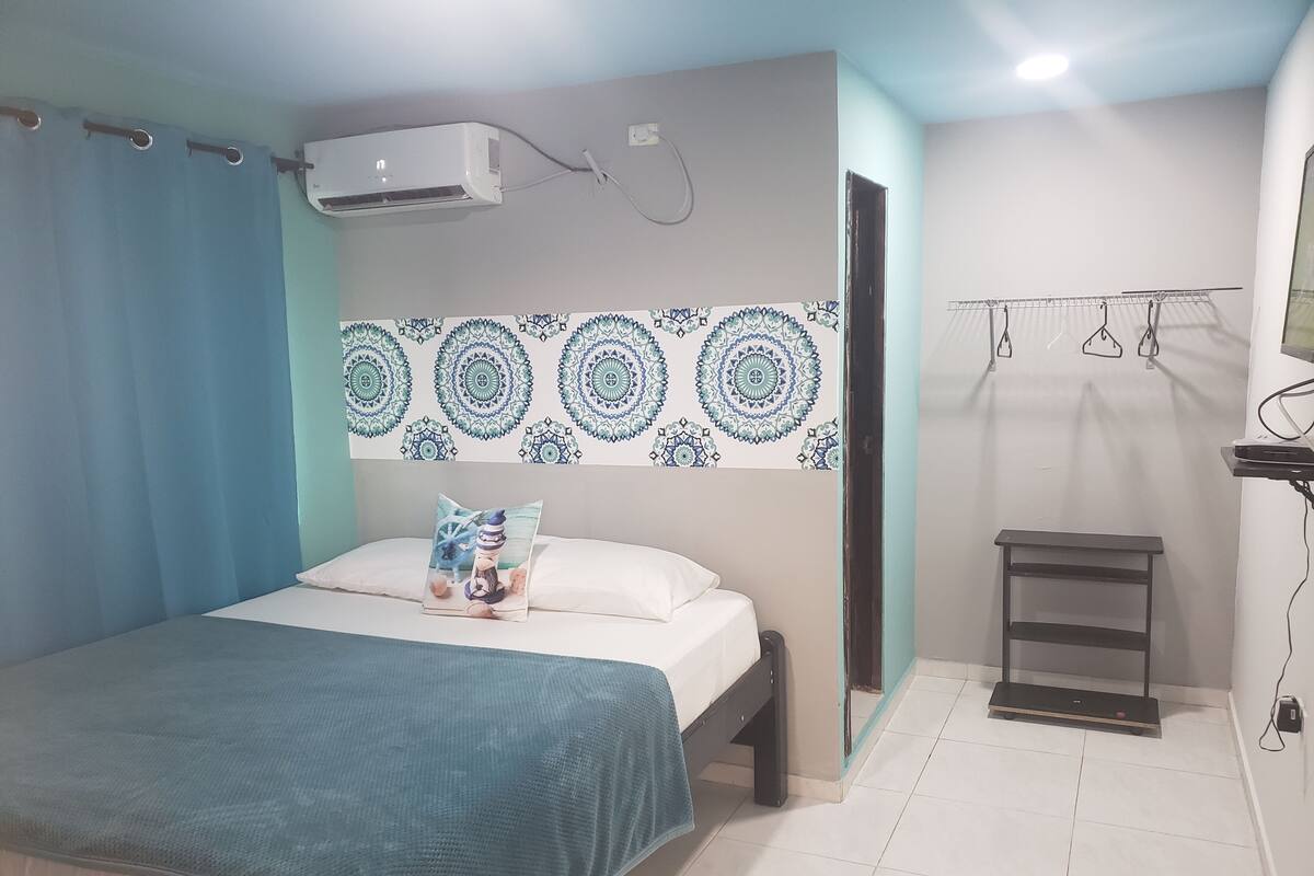 A bedroom with a bed, a wall-mounted air conditioner, a patterned wall decoration, a bedside table, and a hanging clothes rack.