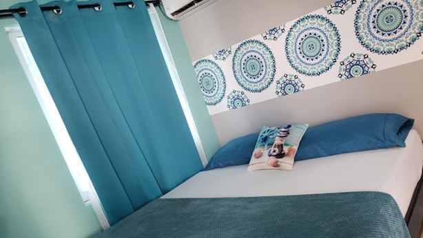 A bed with a blue blanket and pillow, a wall with circular patterns, and a window with blue curtains. - Plaza del Mar (San Andrés)