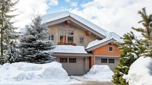 Eine zweistöckige Blockhütte mit Garage, umgeben von schneebedeckten Bäumen und blauem Himmel.