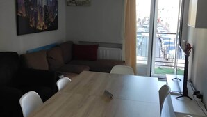 A room with a wooden table, white chairs, a brown sofa, a cityscape painting, an air conditioning unit, and a view of a street through a glass door. - Barajas Apartment. Airport/IFEMA (Madrid)