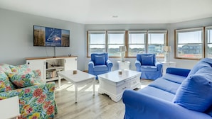 A spacious living room with a large window, a flat-screen TV mounted on the wall, and a floral sofa. - Pelican's Perch | 320 ft from the beach | Private Pool, Hot Tub | Corolla (Corolla)