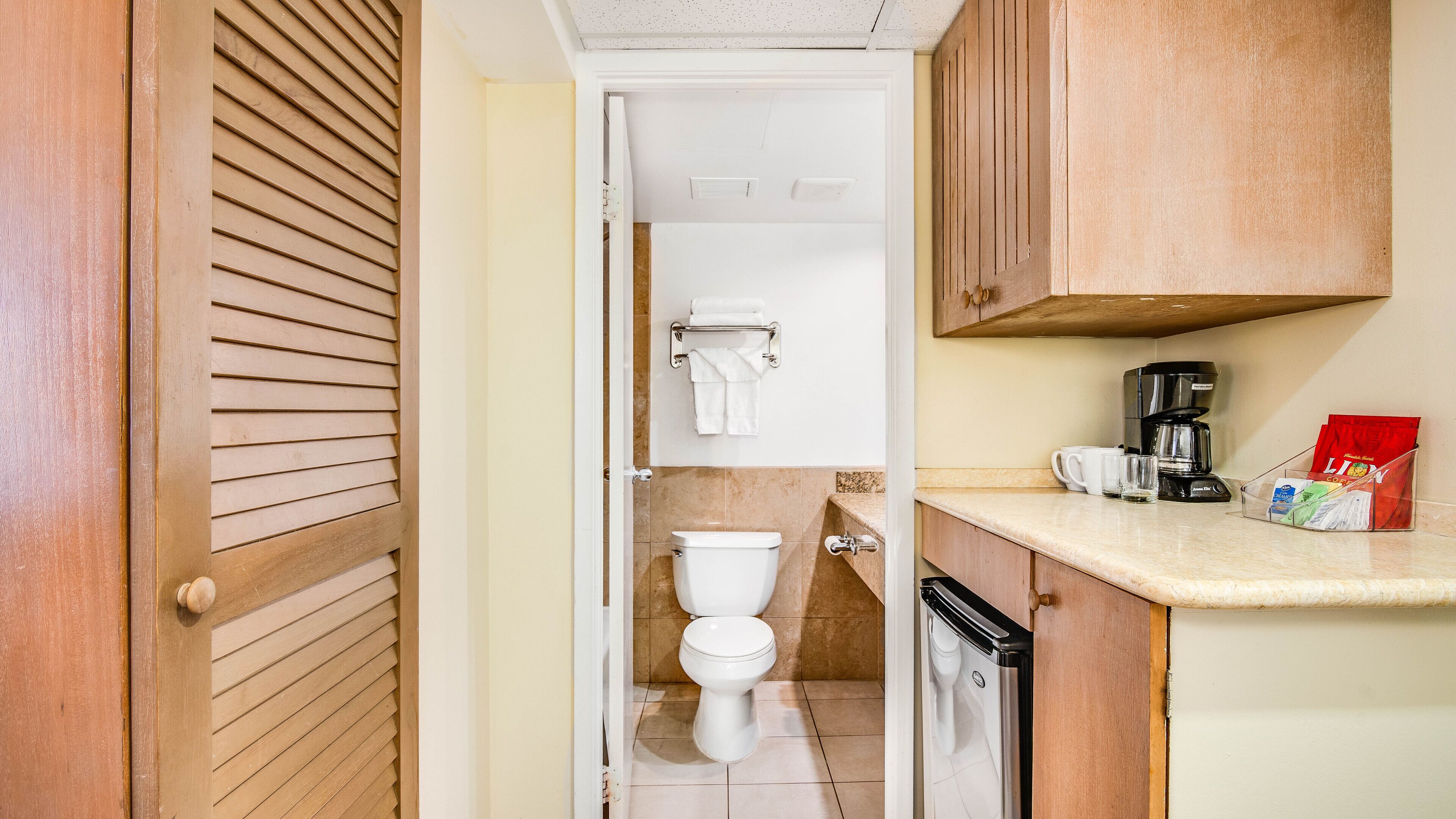 A narrow hallway leading to a bathroom with a toilet, a coffee maker, and a small cabinet with a coffee bag.