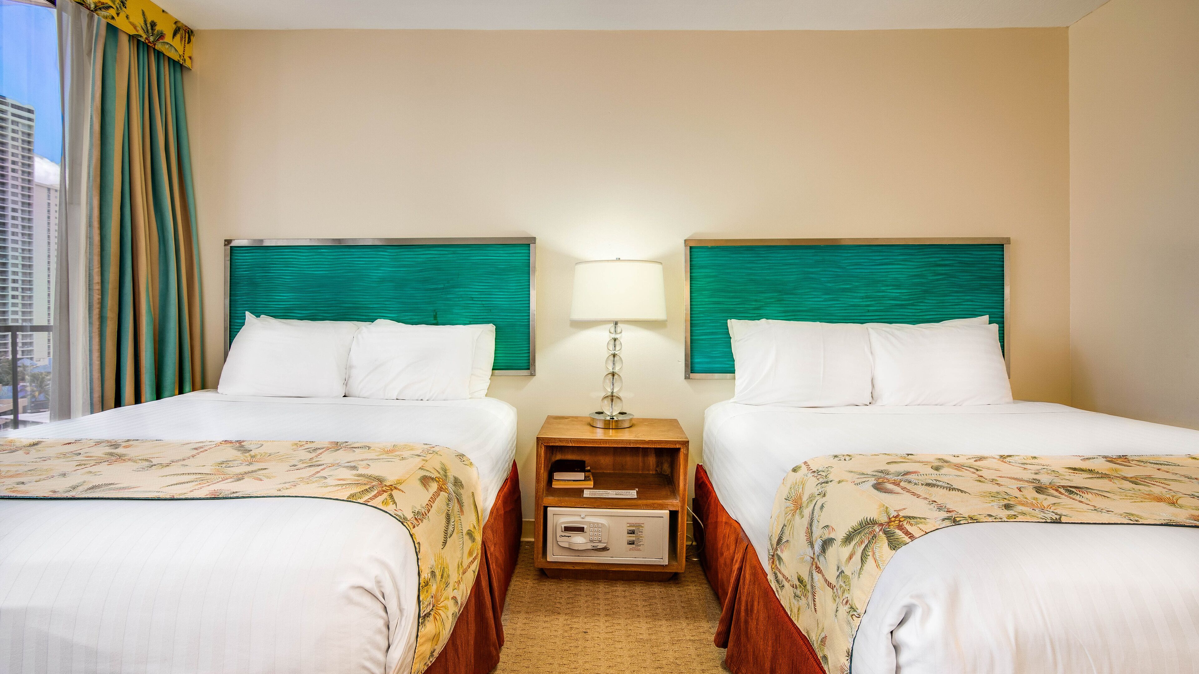 Habitación de hotel con dos camas, cada una con sábanas blancas y colcha de estampado floral.  Entre las camas hay una mesita de noche con una lámpara y un pequeño armario de madera. La habitación tiene vista a un paisaje urbano a través de una ventana con cortinas.