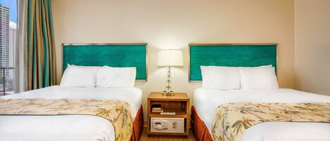 Habitación de hotel con dos camas, cada una con sábanas blancas y colcha de estampado floral. Entre las camas hay una mesita de noche con una lámpara y un pequeño armario de madera. La habitación tiene vista a un paisaje urbano a través de una ventana con cortinas.