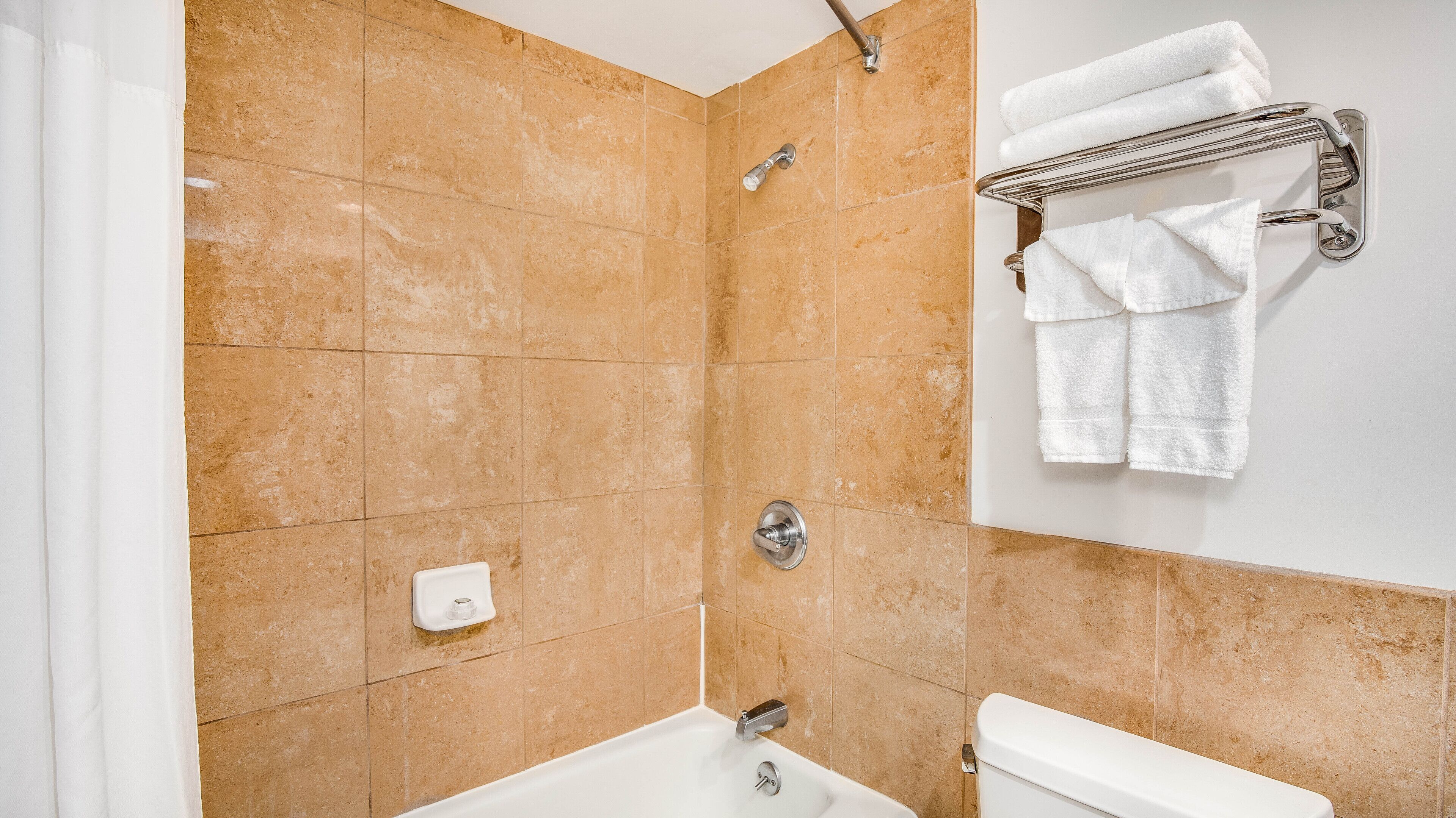 A bathroom with beige tiles, a bathtub, a toilet, a towel rack with towels, and a showerhead.