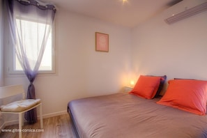 Un dormitorio con cama, mesita de noche, silla, ventana con cortinas translúcidas y aire acondicionado de pared.