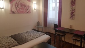Habitación de hotel con cama, escritorio y una silla. Se ve un ventanal con cortinas, una lámpara de pared y un cuadro de una rosa.