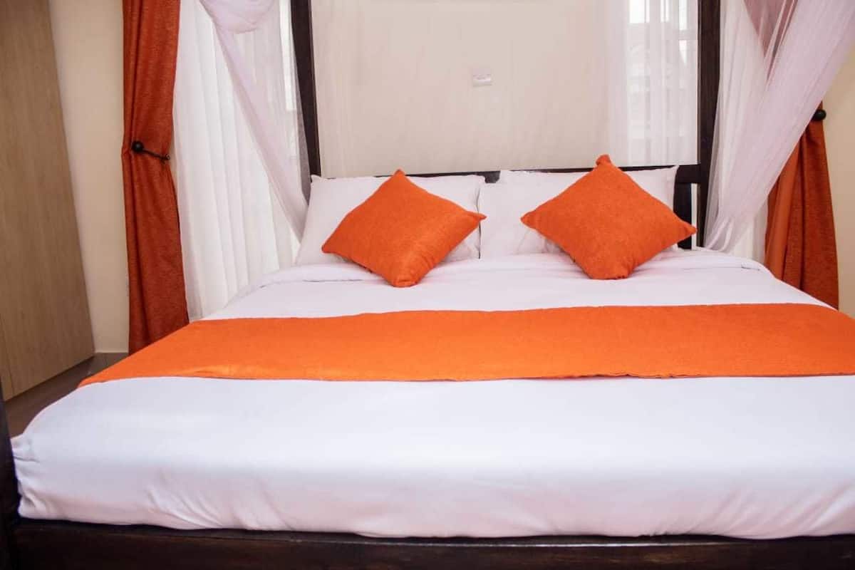 A bed with white bedding and orange accents, a wooden headboard, and sheer curtains.