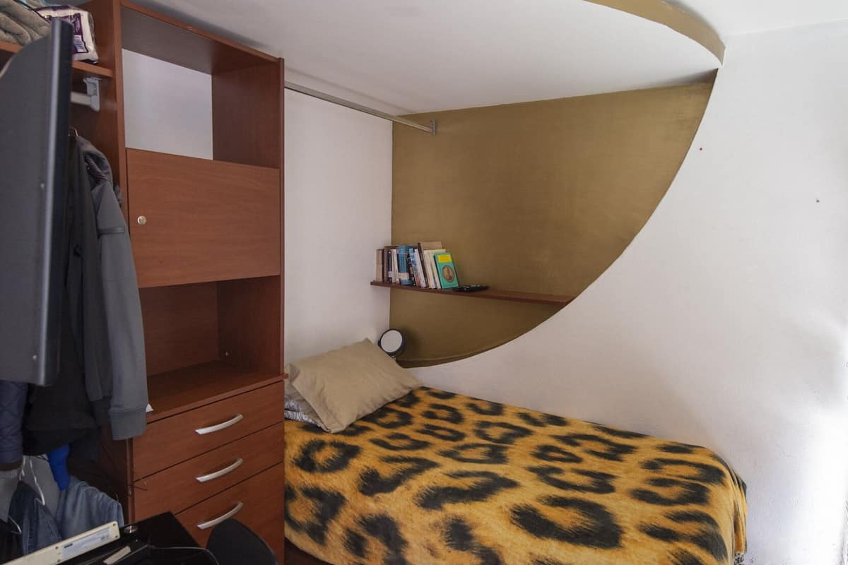 A compact room with a curved ceiling, a bed with a leopard print cover, a wooden wardrobe, a desk with a laptop, and a bookshelf.