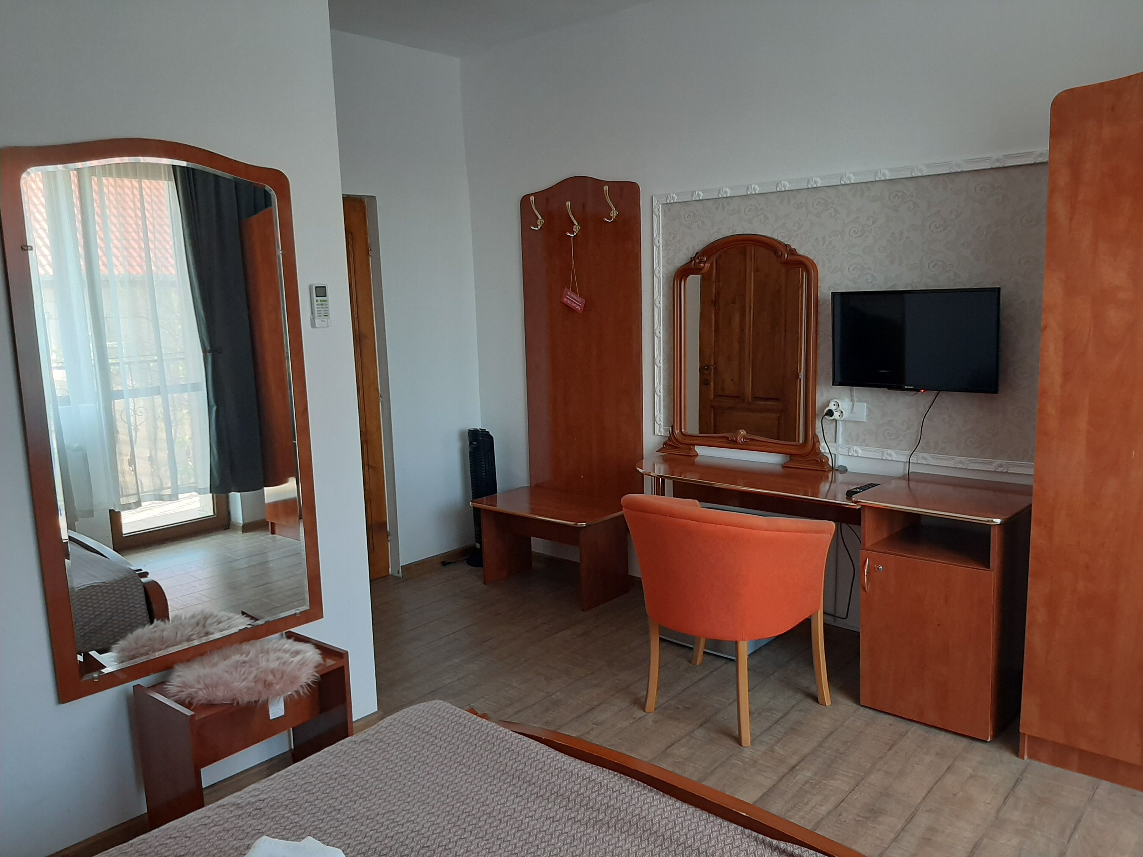 Ein Zimmer mit Schreibtisch aus Holz, einem Stuhl, einem Spiegel, einem an der Wand befestigten Fernseher und einem Bett.