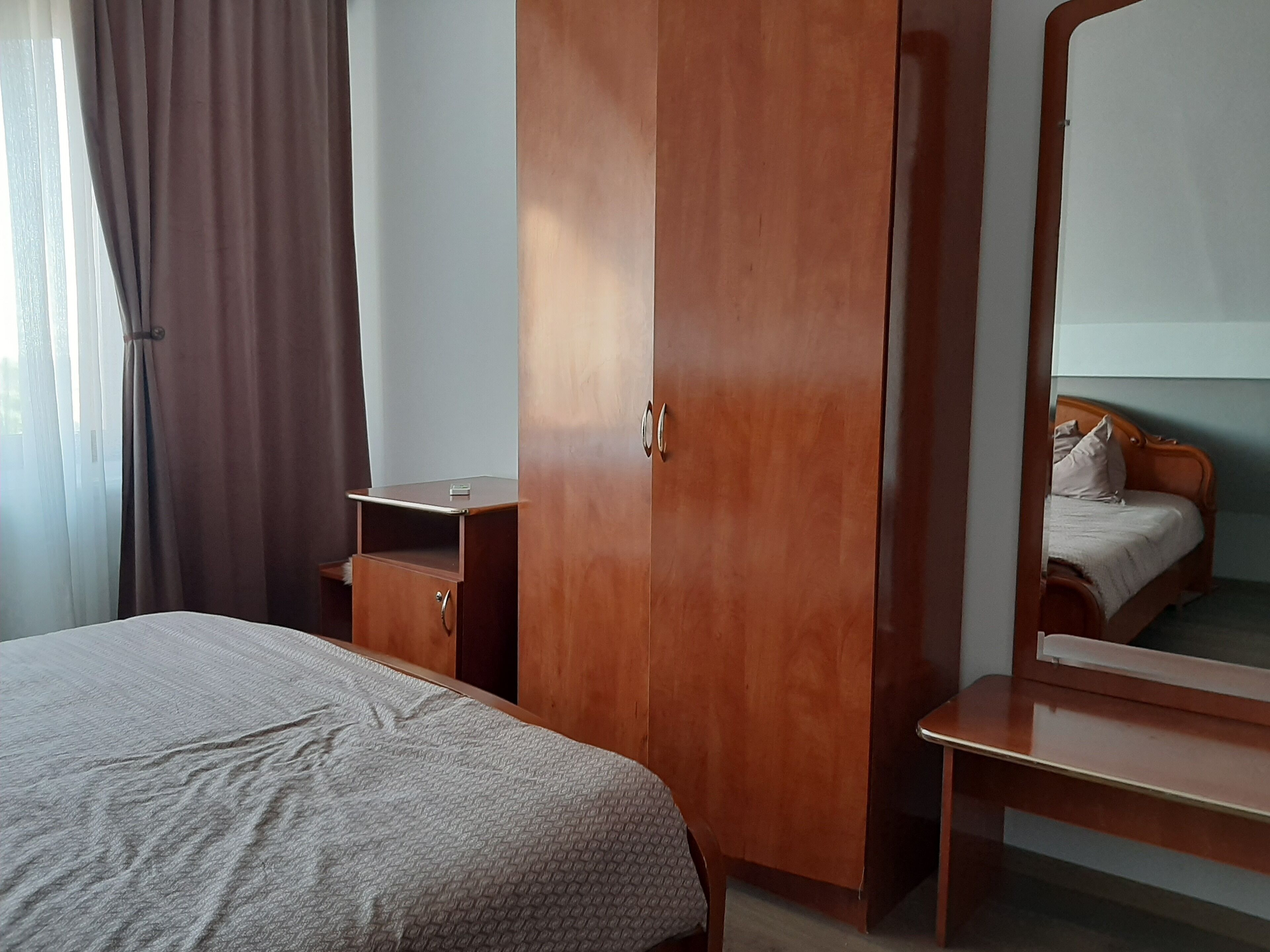 A bedroom with a bed, a wooden wardrobe, a bedside table, and a mirror reflecting a bed.