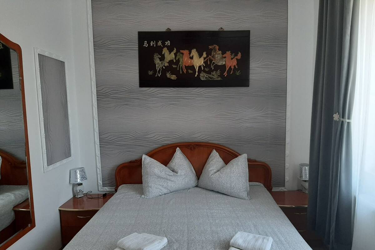 Ein ordentlich bezogenes Bett mit Kissen und gefalteten Handtüchern, ein hölzerner Kopfteil, ein Wandbild und ein Spiegel.