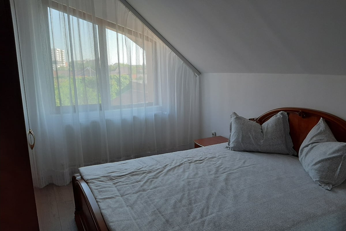 Ein Schlafzimmer mit einem Bett, einem Nachttisch und einem Fenster mit transparenten Vorhängen.