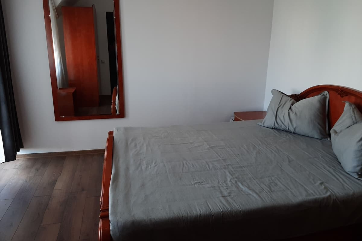 Ein Schlafzimmer mit einem hölzernen Bett, grauer Bettwäsche, einem Nachttisch aus Holz, einem Spiegel und einem Holzboden.