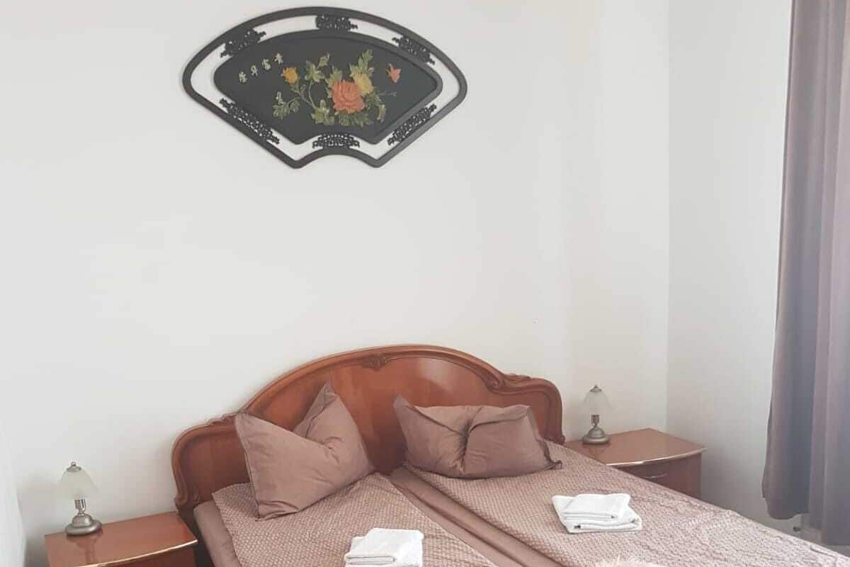 Ein Doppelbett mit Holz-Kopfteil, zwei Nachttische mit Lampen, ein Wanddekor und ein Fenster mit Vorhängen.