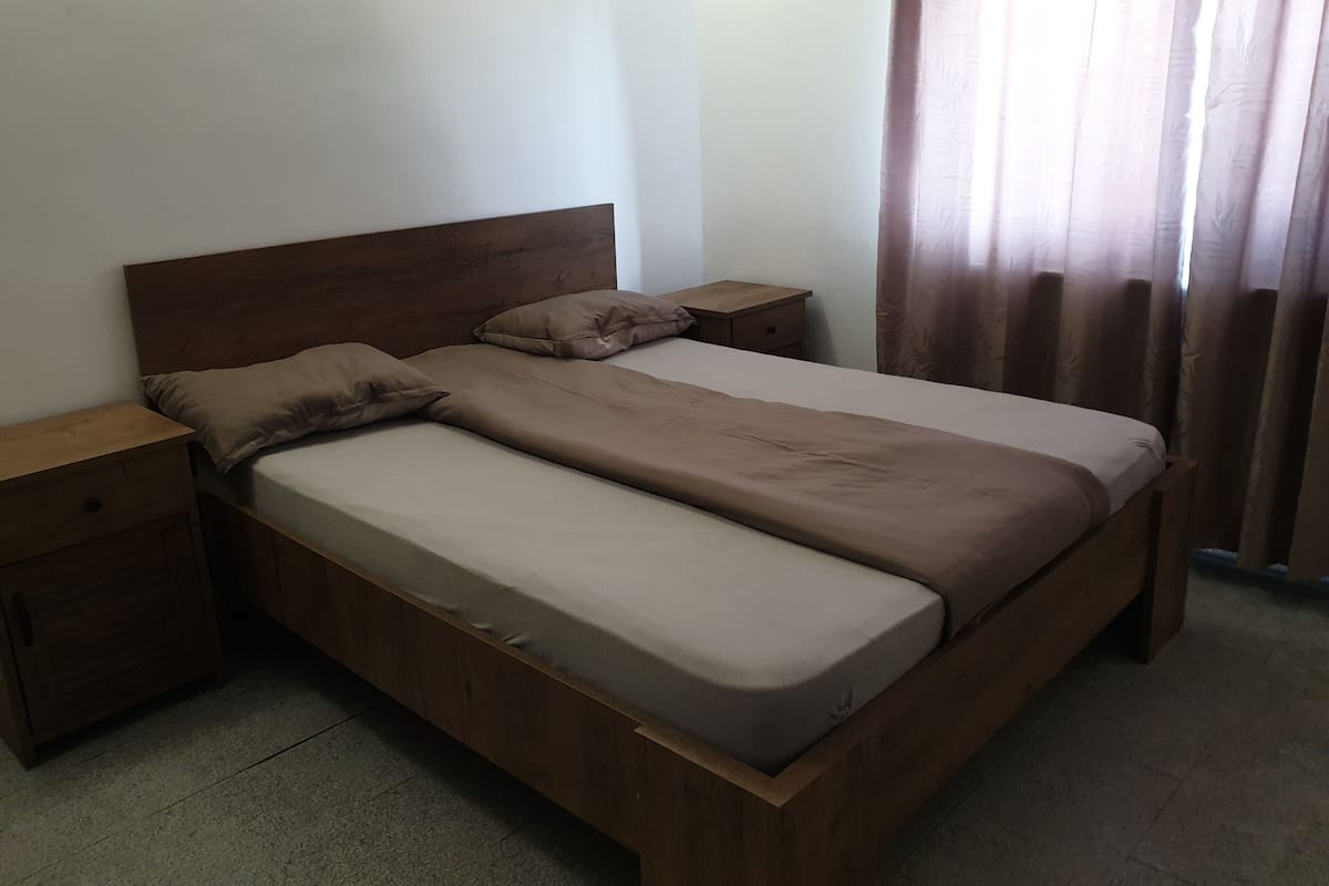 Ein Zimmer mit einem Einzelbett, einem Holzkopfende, zwei Nachttischen und einem Vorhangfenster.